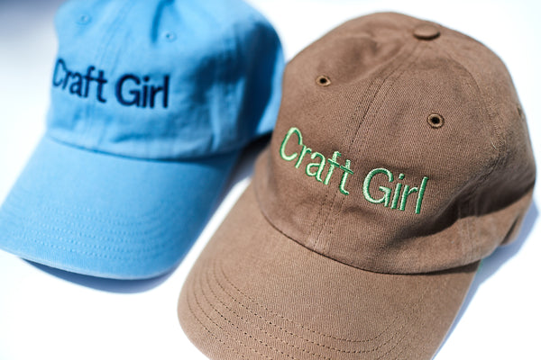 Blue and brown 6-panel baseball caps with the text Craft Girl embroidered on the front panel in a san serif typeface. 